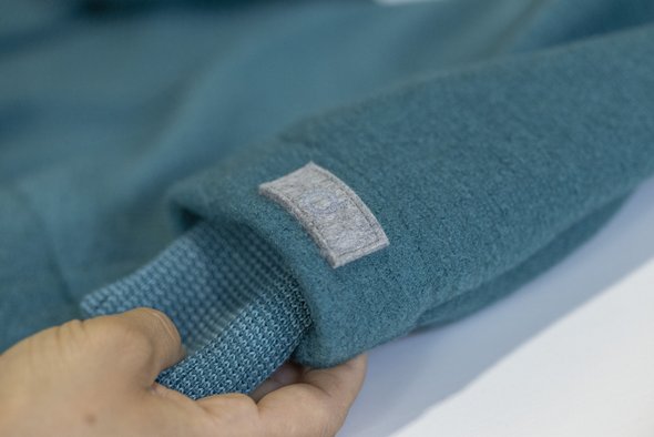 Eine Hand hält das Strick-Bündchen in Pacific-Lagoon an einer pazifikblauen disana Outdoor-Jacke aus Wollwalk.