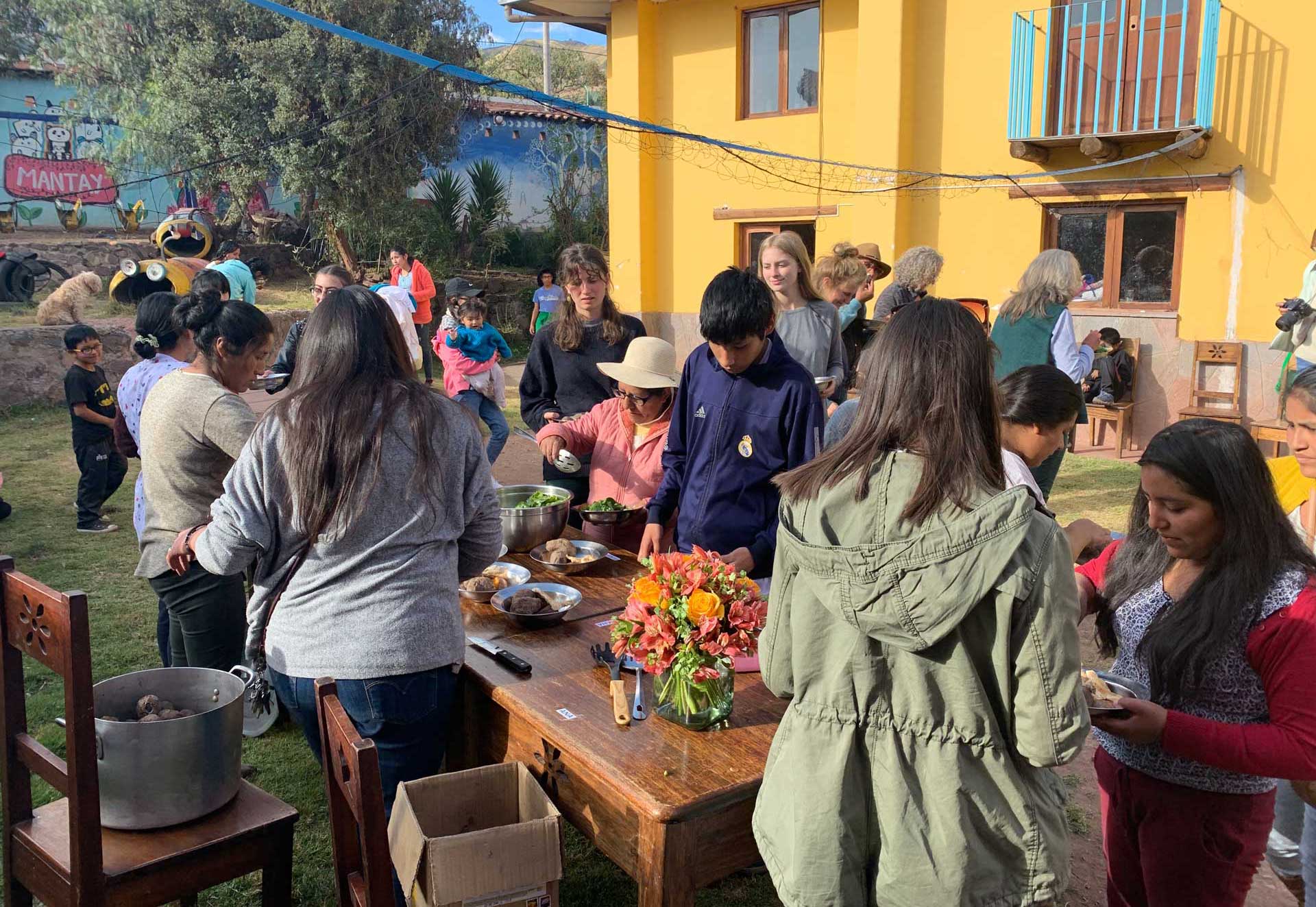 Casa Mantay, Cusco Peru: Peruaner bei der Essensausgabe an einem Holztisch mit orangem Blumenstrauß vor einem gelben Gebäude.
