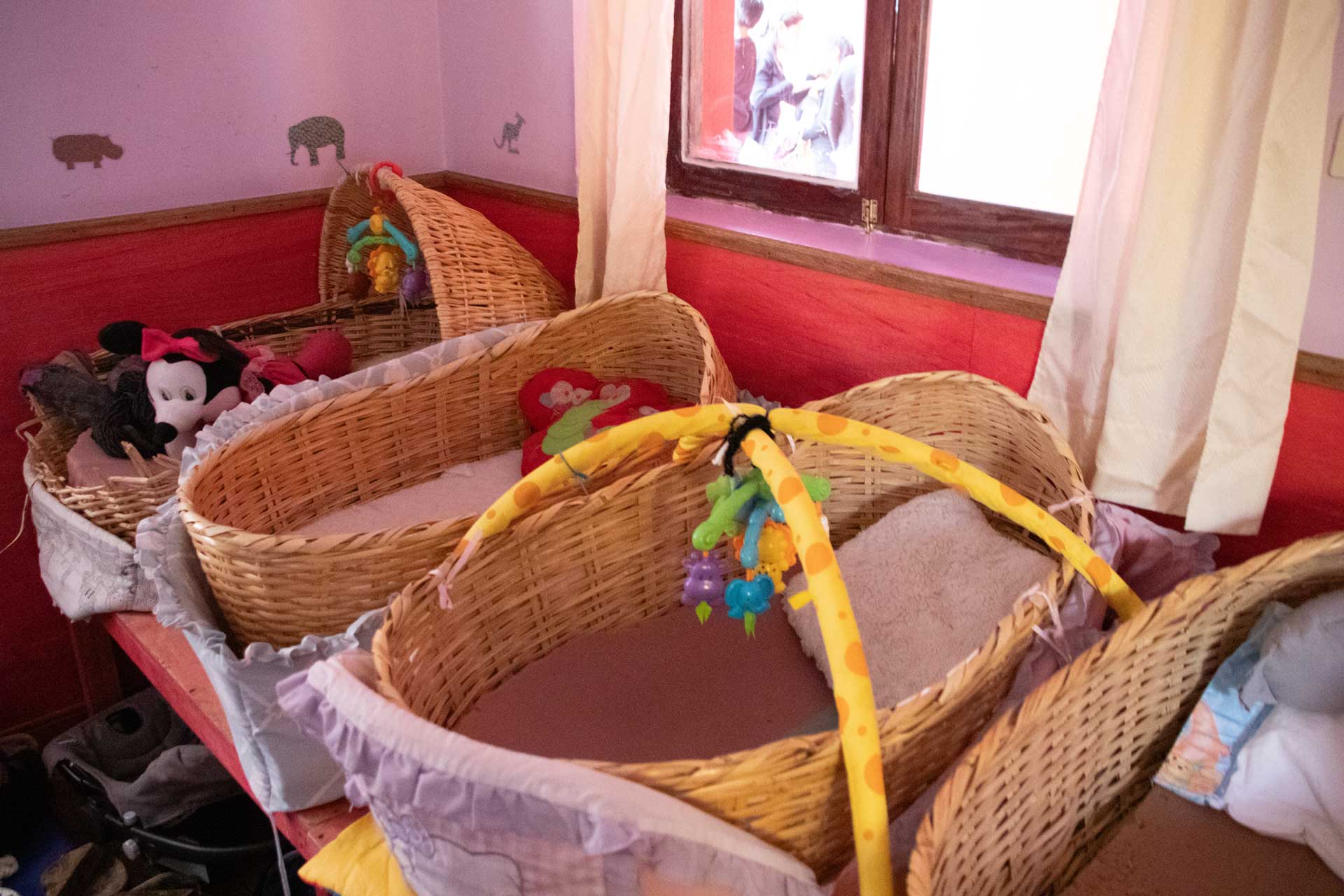 Casa Mantay, Cusco, Peru: Mehrere geflochtene Weidenkörbe als Babybetten mit Spieltrapez & Kuscheltieren vor einer roten Wand
