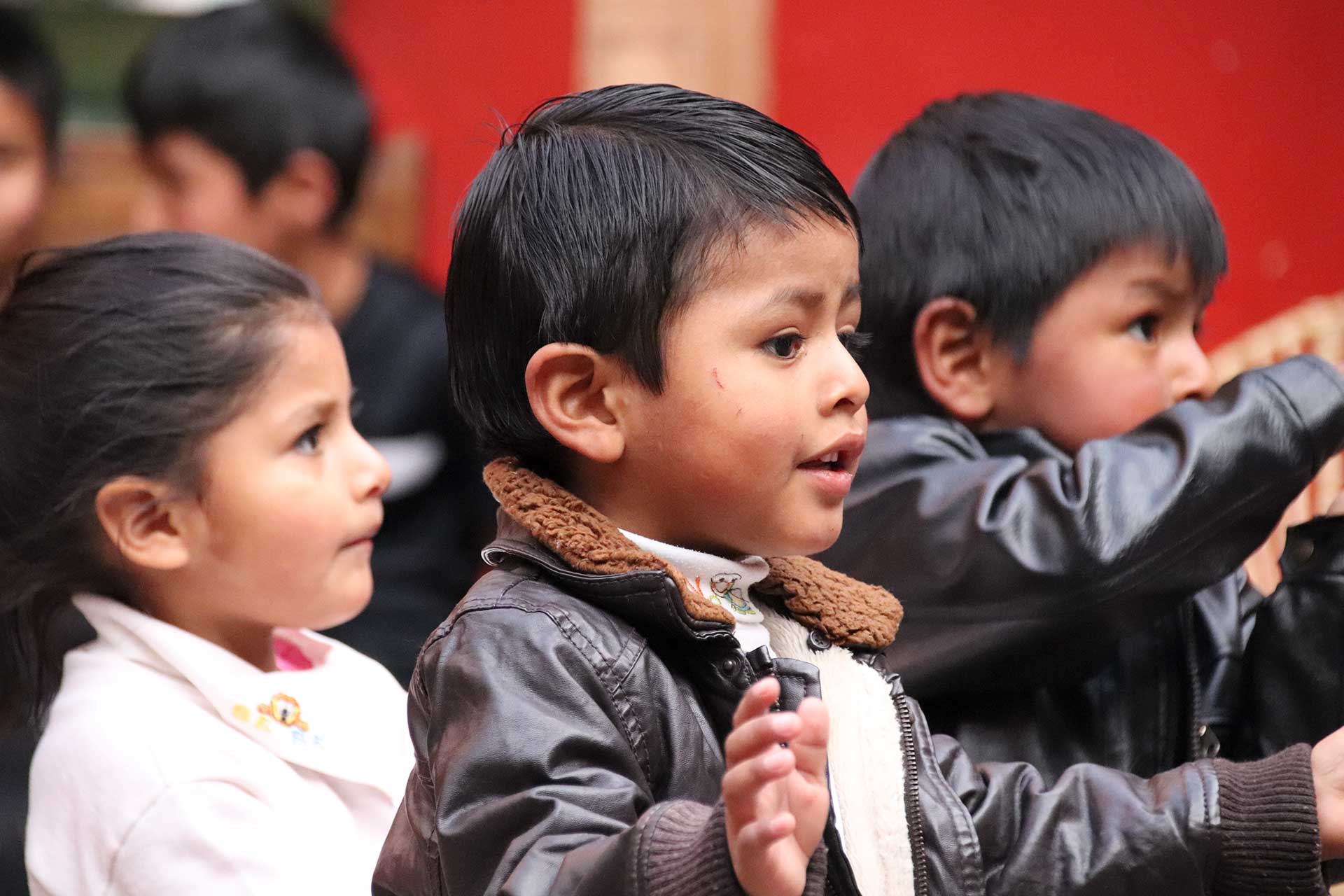 Casa Mantay Cusco Peru: 3 Kinder blicken aufmerksam nach rechts, die Jungen tragen dunkle Lederjacken: vor roter Wand
