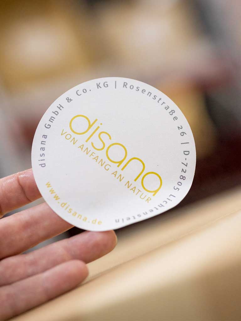 Rundes disana Logo-Etikett für den Paketversand mit Adresse in Grau, Slogan ‚Von Anfang an Natur in Gold, weißer Hintergrund