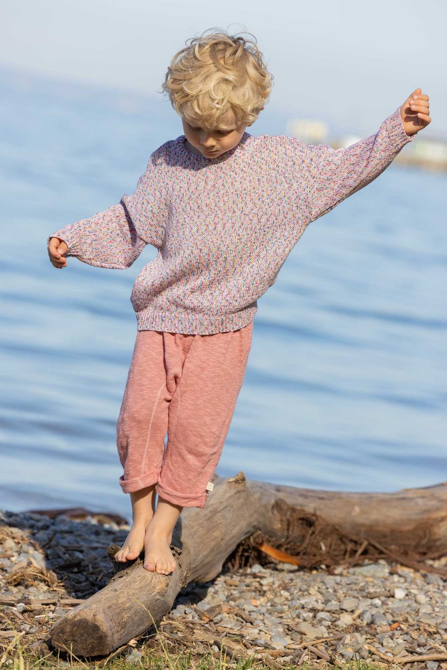 Junge in disana Baumwoll-Pullover "Spray", Softpink Spray & Leinen-Strickhose, Softpink, balanciert auf Holzstamm am Seeufer