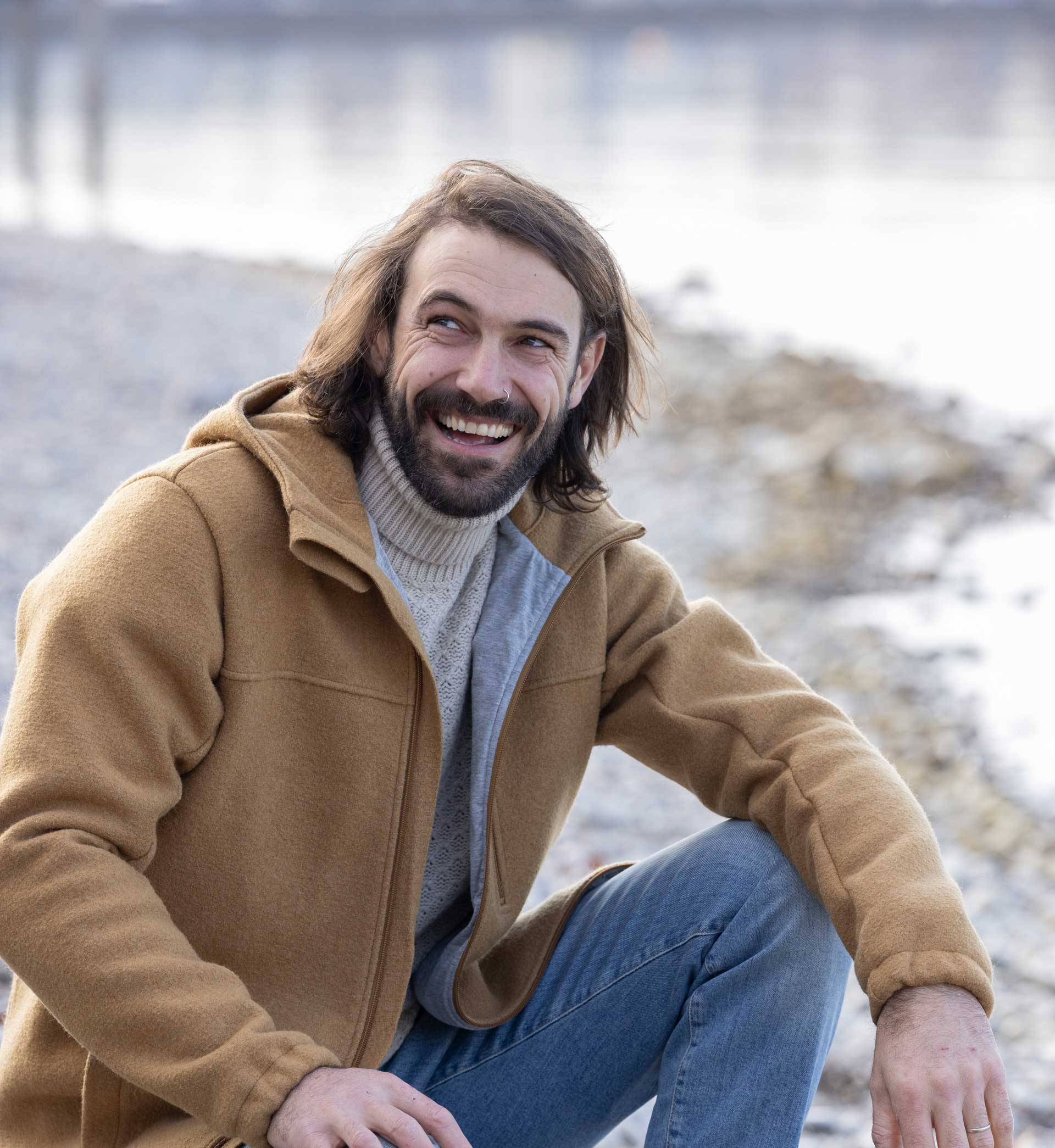 Lachender braunhaariger Mann in disana Herren-Jacke in Karamell und hellem Rollkragenpullover am kiesigen Ufer eines Sees.