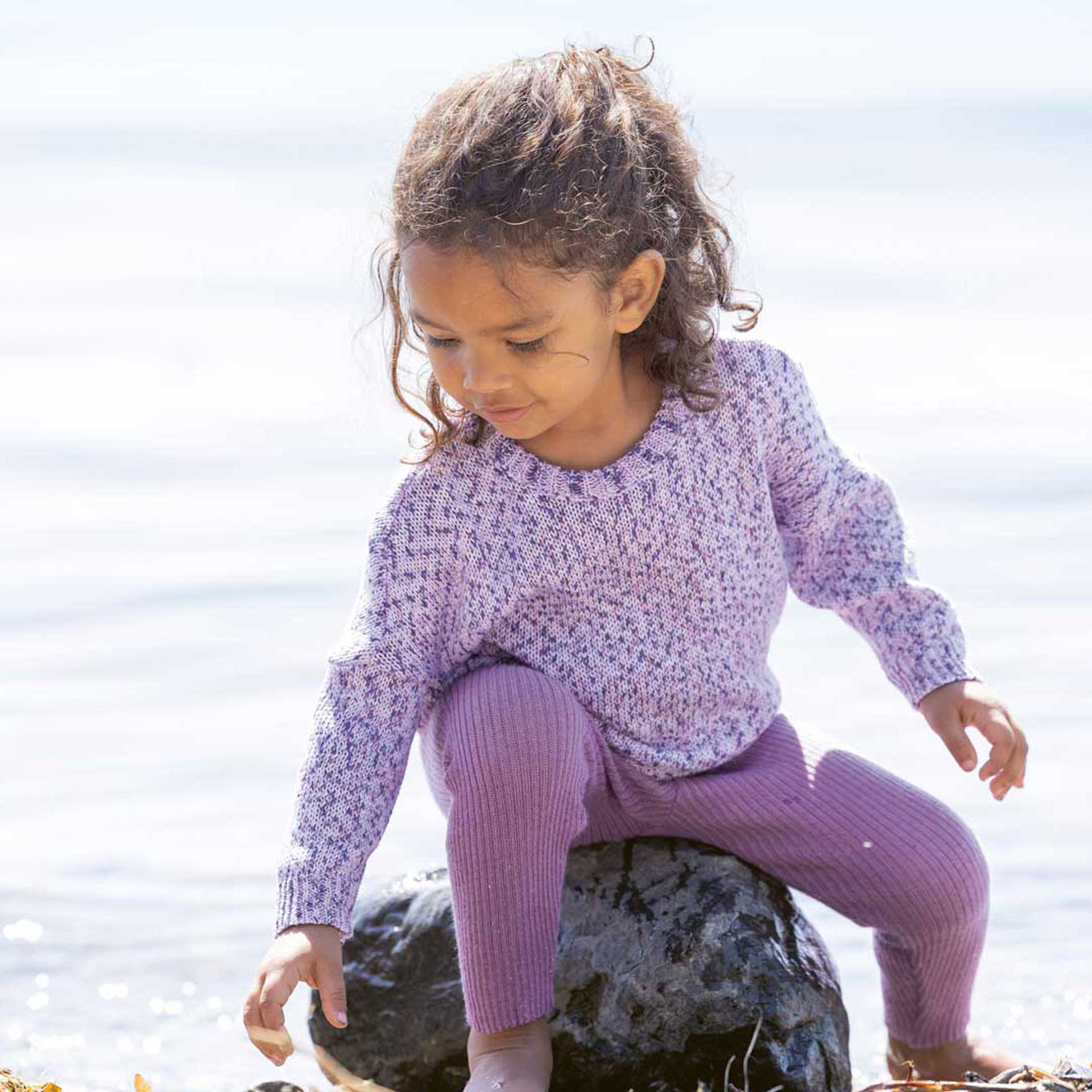 Mädchen mit disana Baumwoll-Pullover "Flieder Spray" und Leggings-Light Flieder schaut nach Muscheln vor Seehintergrund.