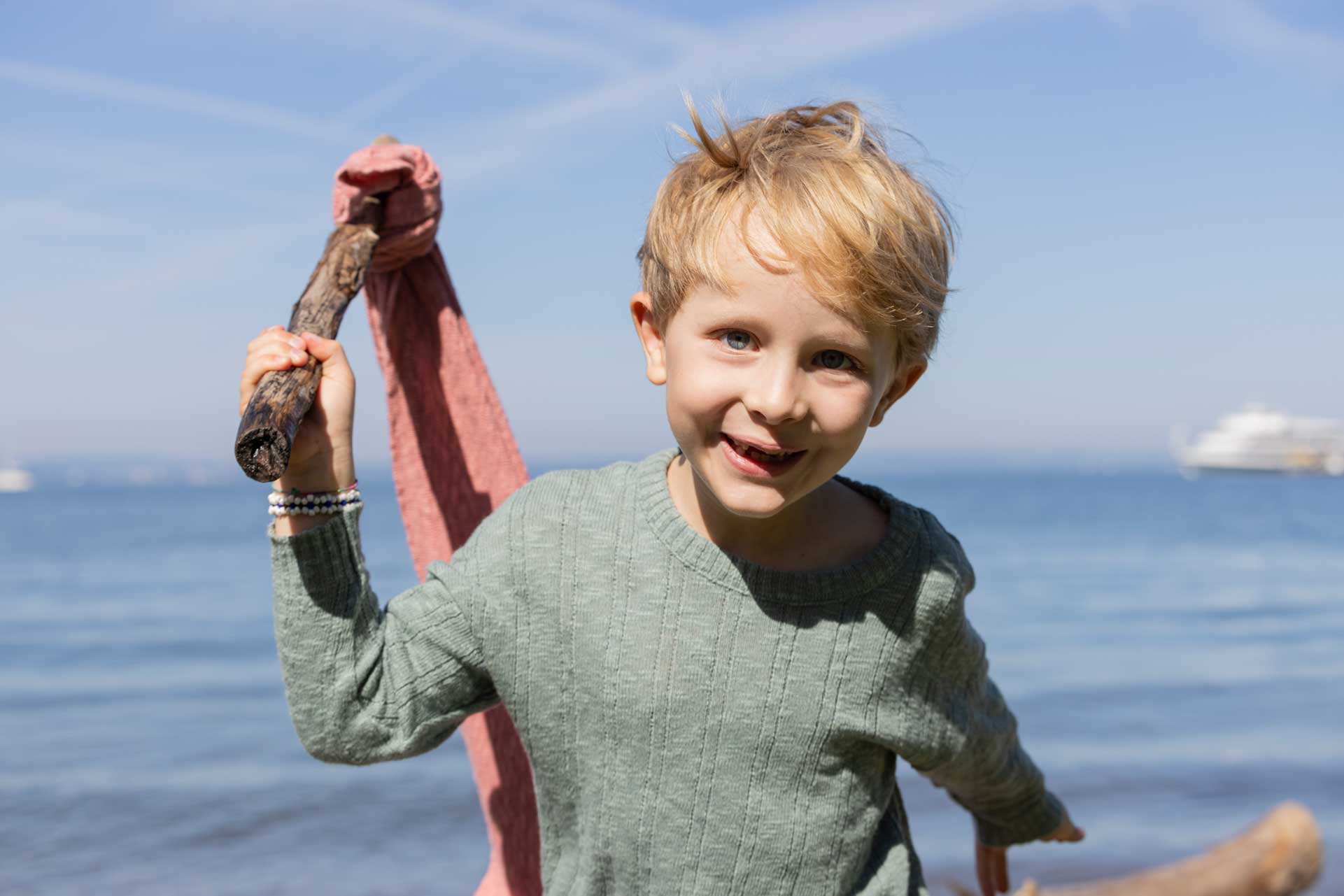 Junge im jadefarbenen disana Leinen-Pullover aus Bio-Baumwolle-Leinen-Mix spielt lachend mit einem Stock am See.