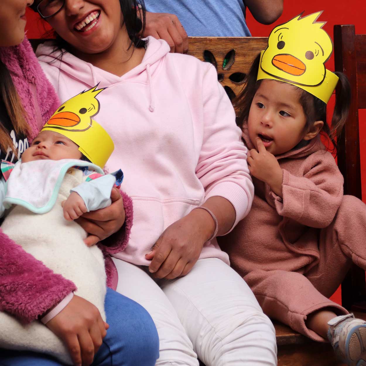 Casa Mantay Cusco Peru: Lachende Frau mit Baby & Kleinkind, die gelbe Enten-Papierkronen tragen, vor einem roten Hintergrund.