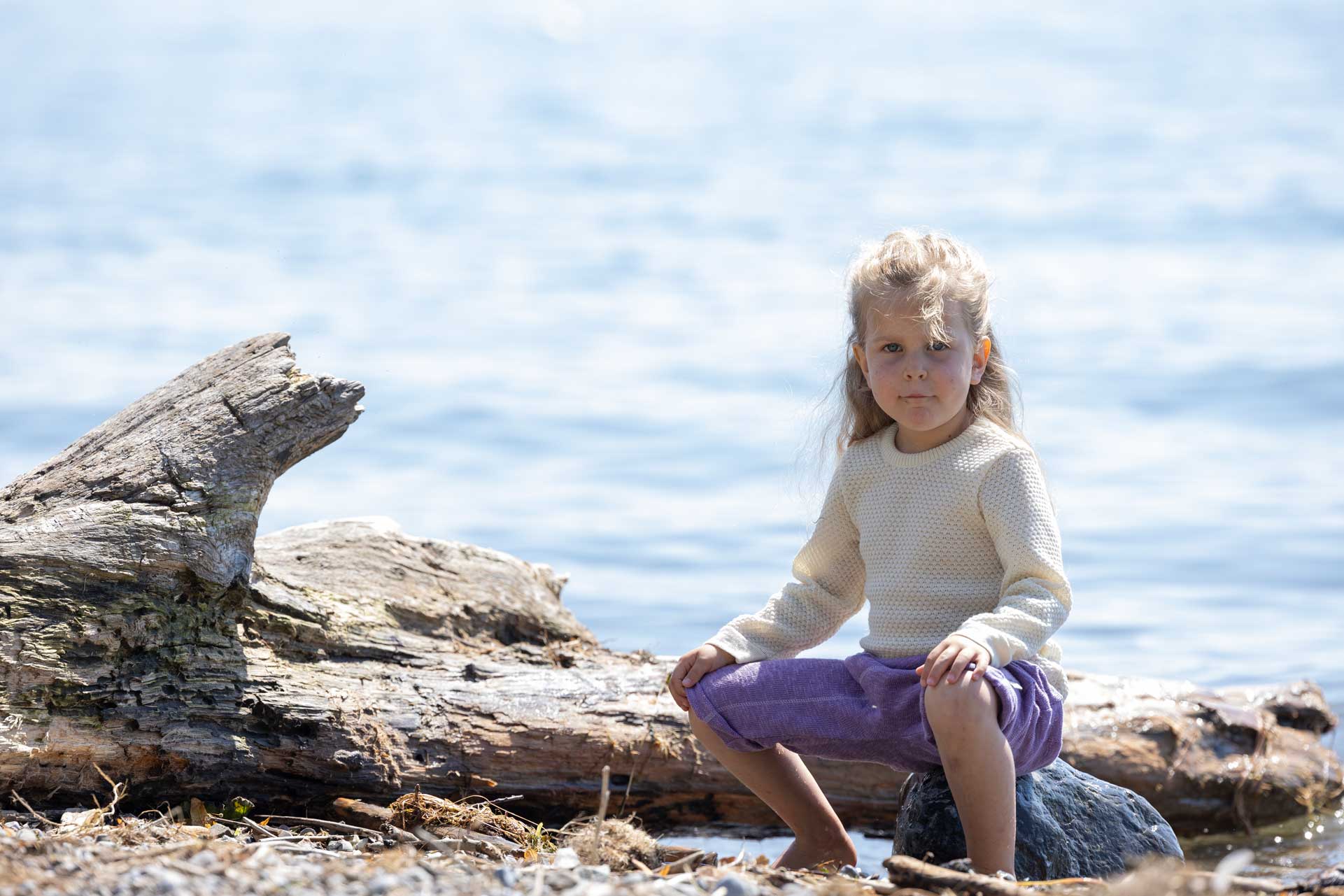 Mädchen im naturfarbenen disana Wabenstrick-Pullover und Leinen-Strickhose in Flieder sitzt auf einem Stein am Seeufer.