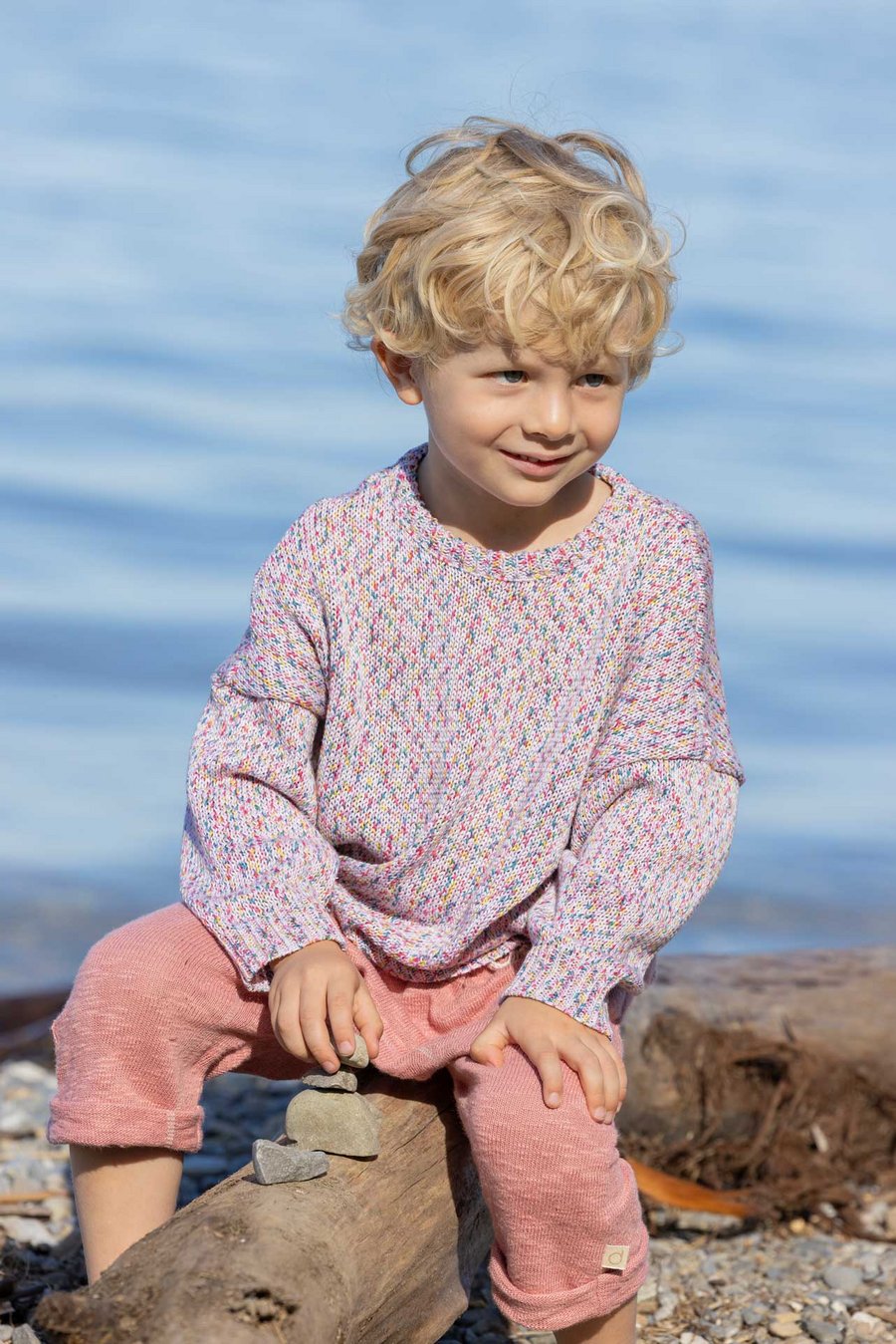 Blondes Kind im disana Baumwoll-Pulli Spray, Softpink Spray & Leinen-Strickhose Softpink spielt auf Holzschaukel am Seeufer