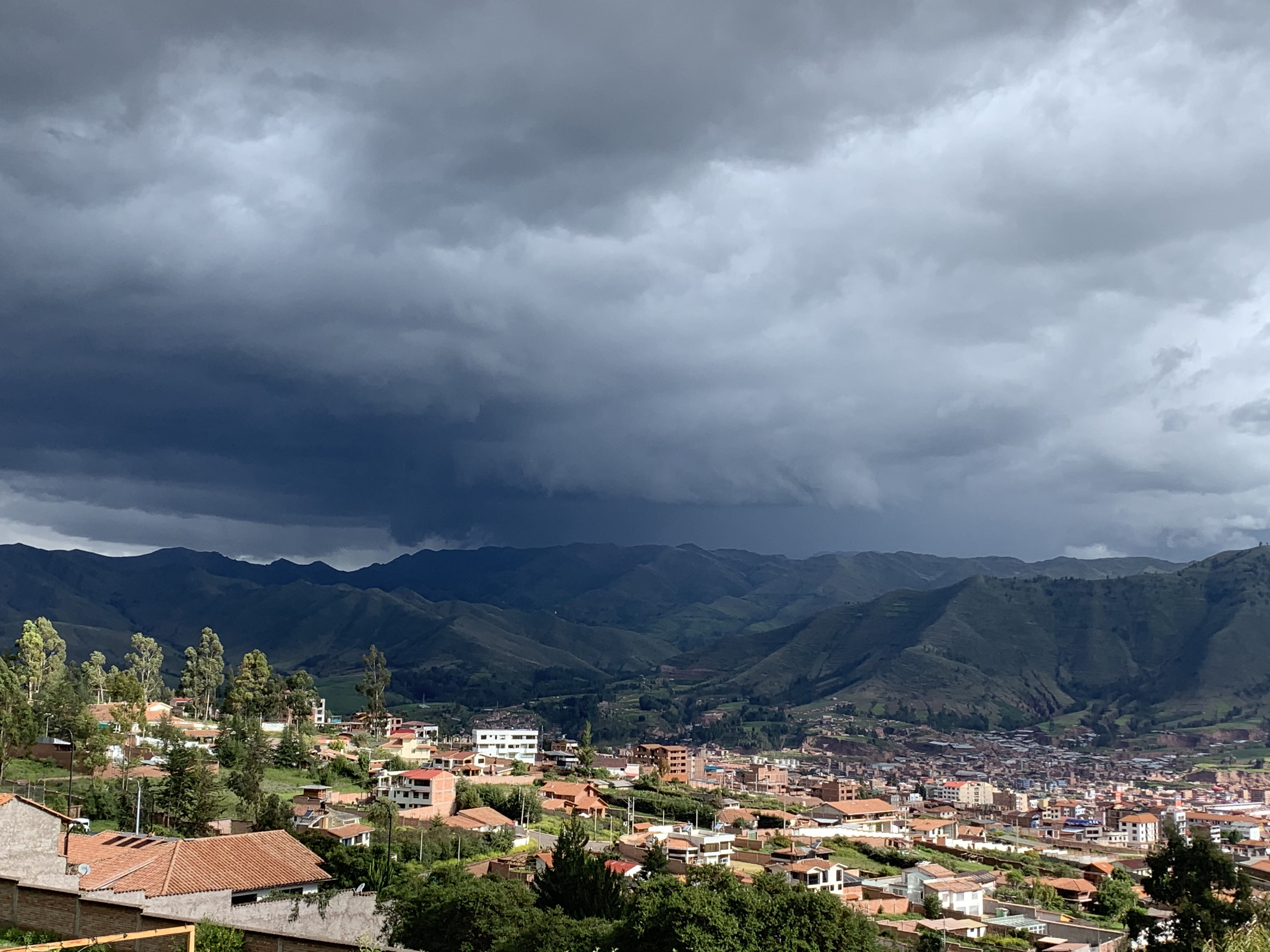 Panorama von Cusco, Peru: Rote Ziegeldächer im Andental vor grünen Bergen unter dunklem Gewitterhimmel.